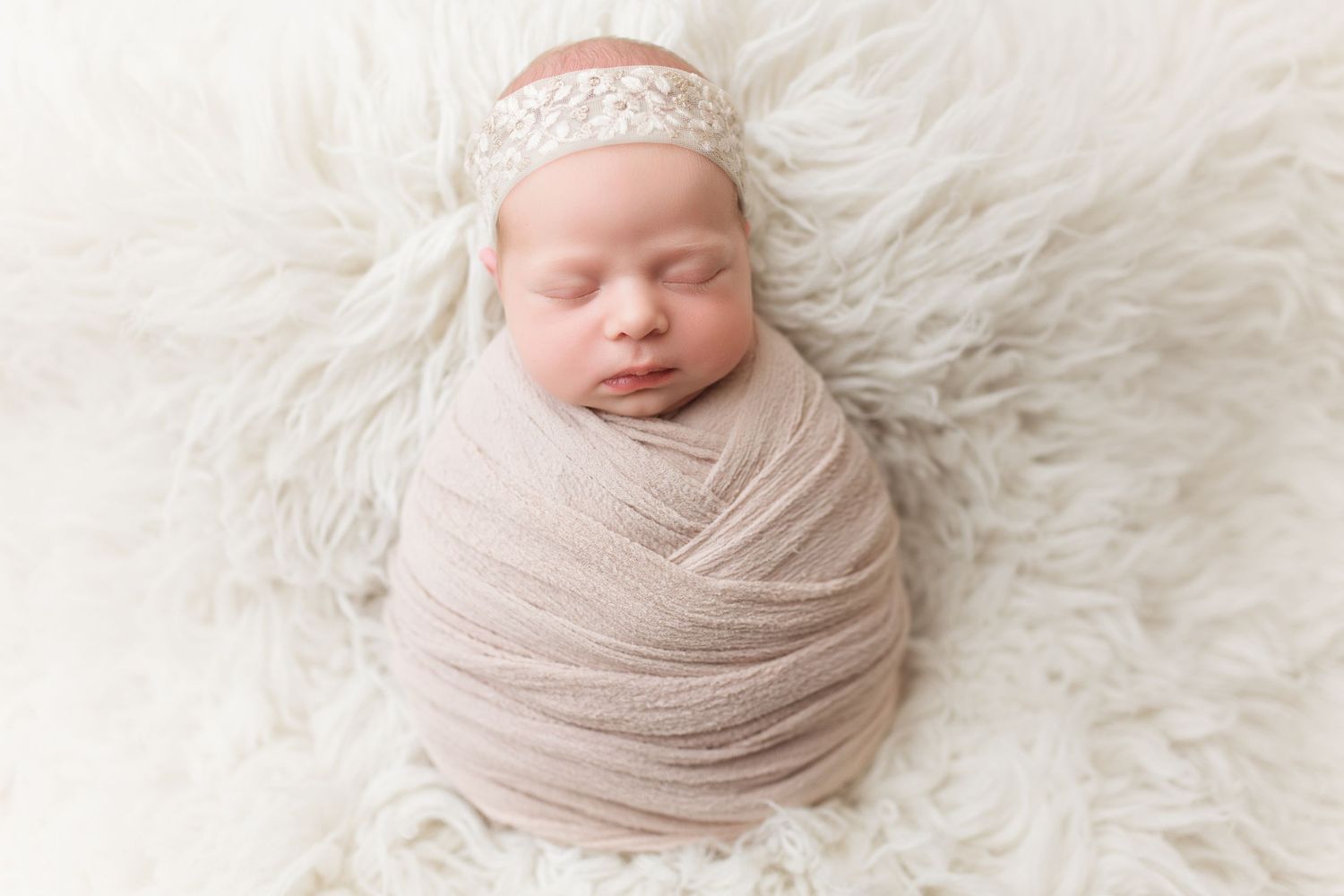 Sleeping newborn wrapped in beige fabric with lace headband on fluffy white fur background.