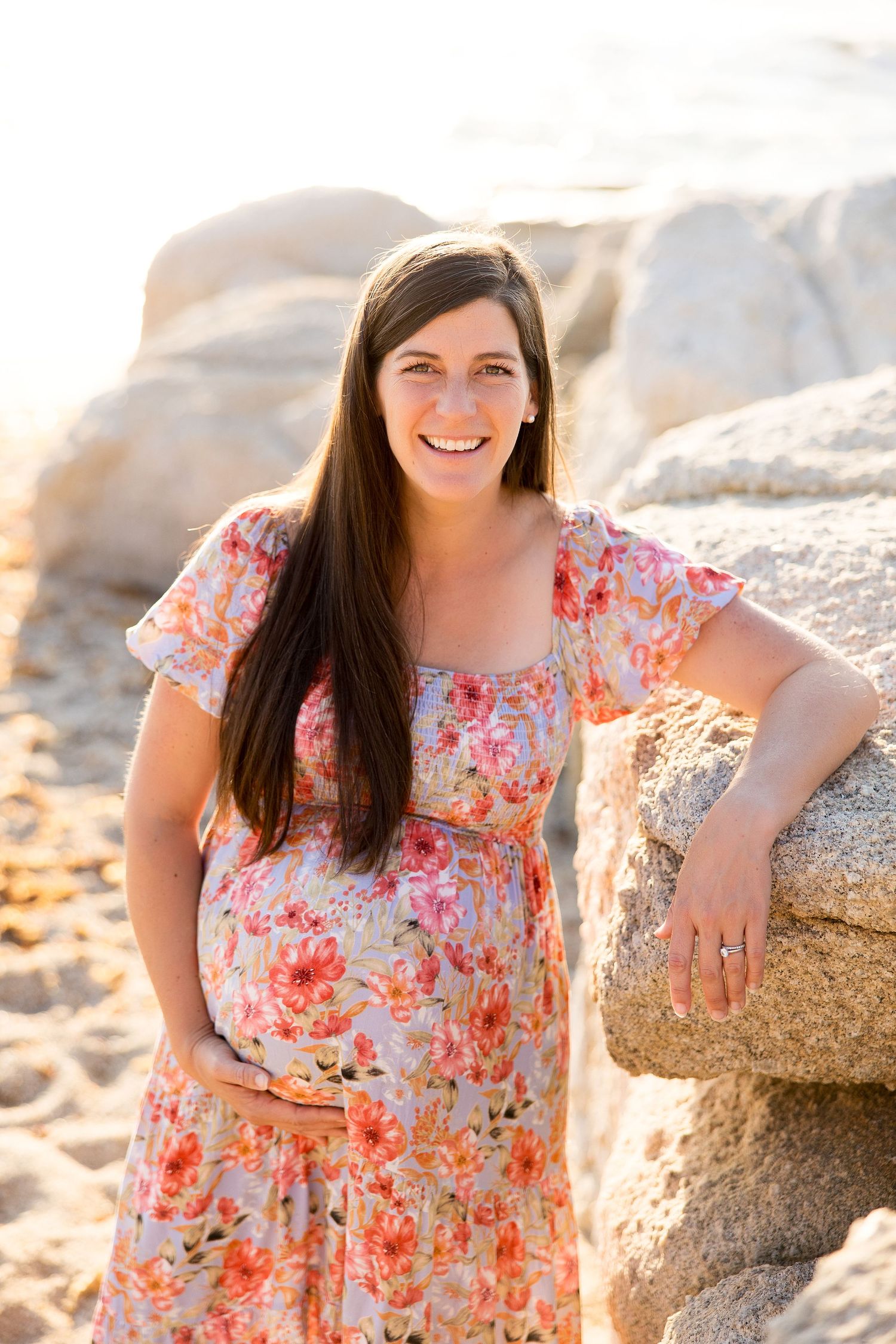 Woman in a floral dress poses against coastal rocks during sunset.