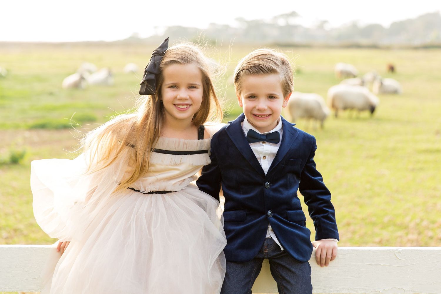Two children in dressy outfits smile while posing near white fence on farm.