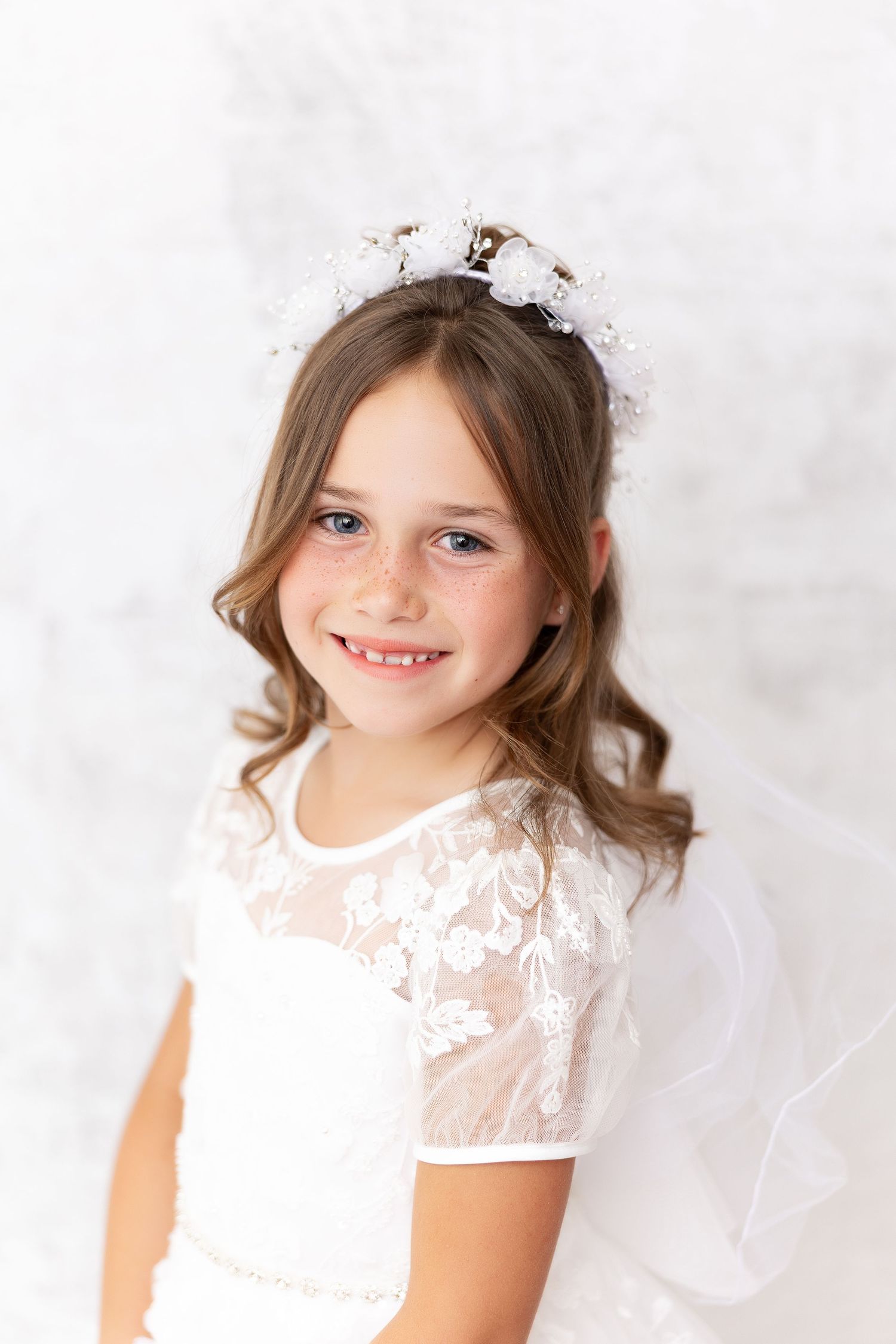 Child wearing white lace dress with delicate floral details and sheer sleeves poses against light background.