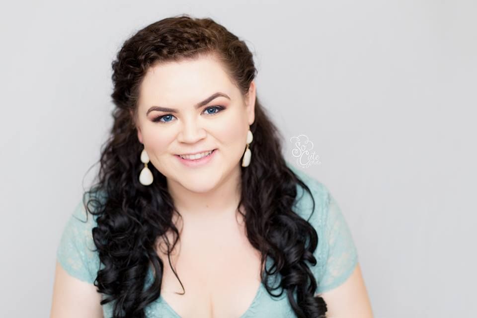 A person with long dark curly hair wearing a mint-colored top and pearl drop earrings smiles against a light gray background.