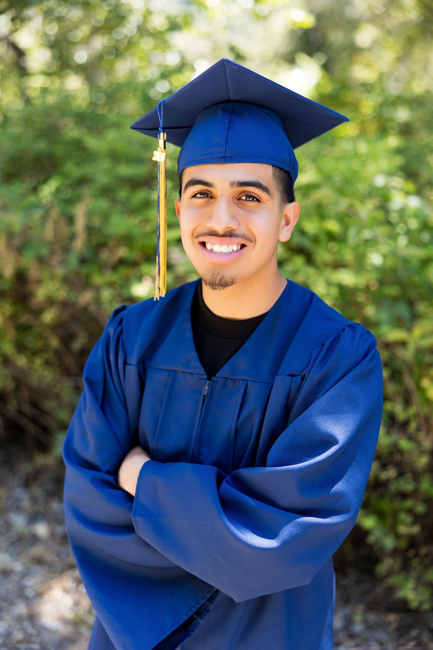 Smiling young man in blue graduation cap and gown with arms crossed outdoors.