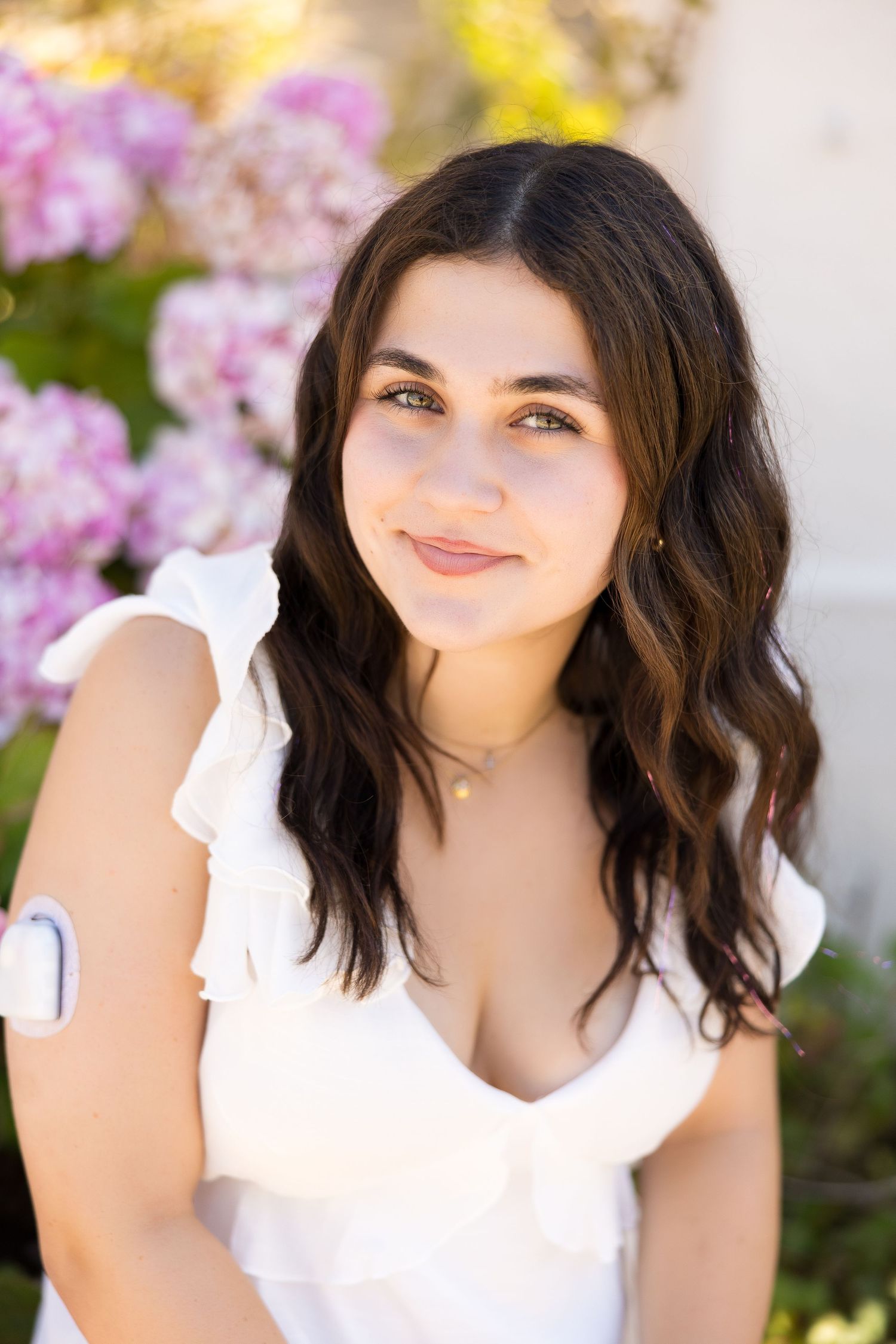Young woman with dark wavy hair in white ruffled top smiling outdoors with pink hydrangeas in background.