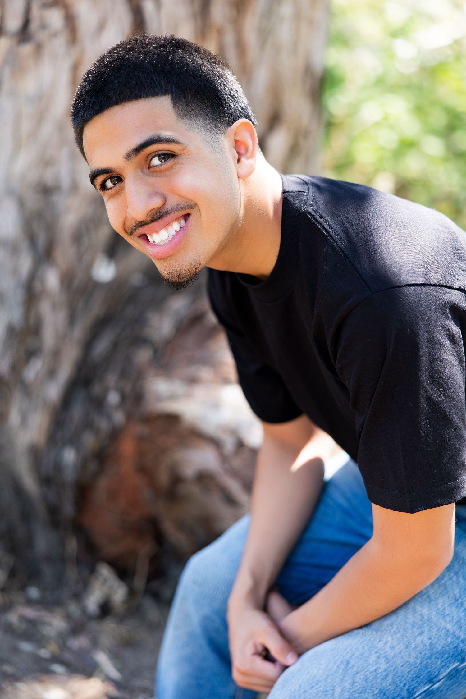 High school senior smiling  in black t-shirt and jeans sitting in Carmel Valley during his senior portrait session.