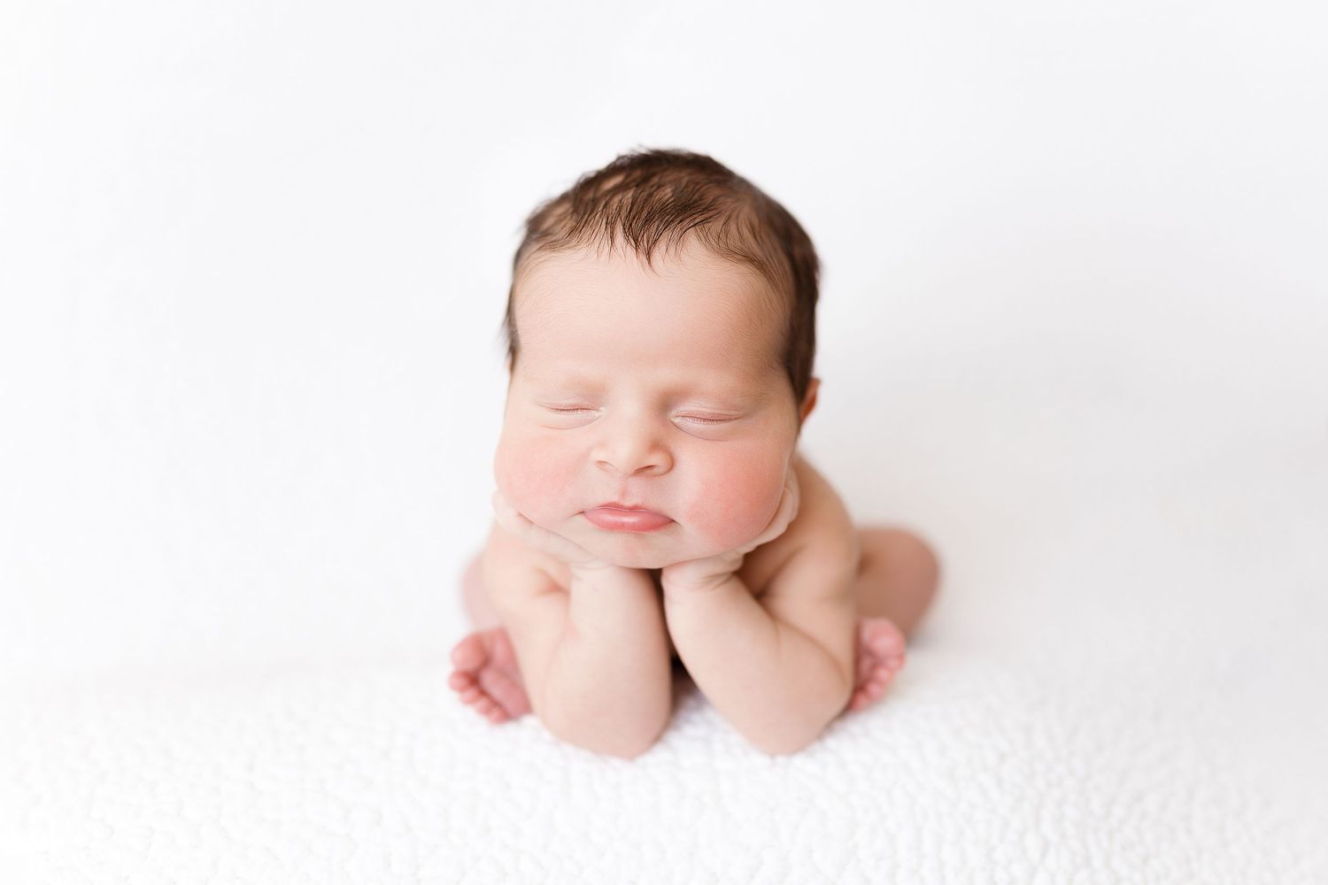 Newborn baby posed with hands under chin on white backdrop in studio setting.