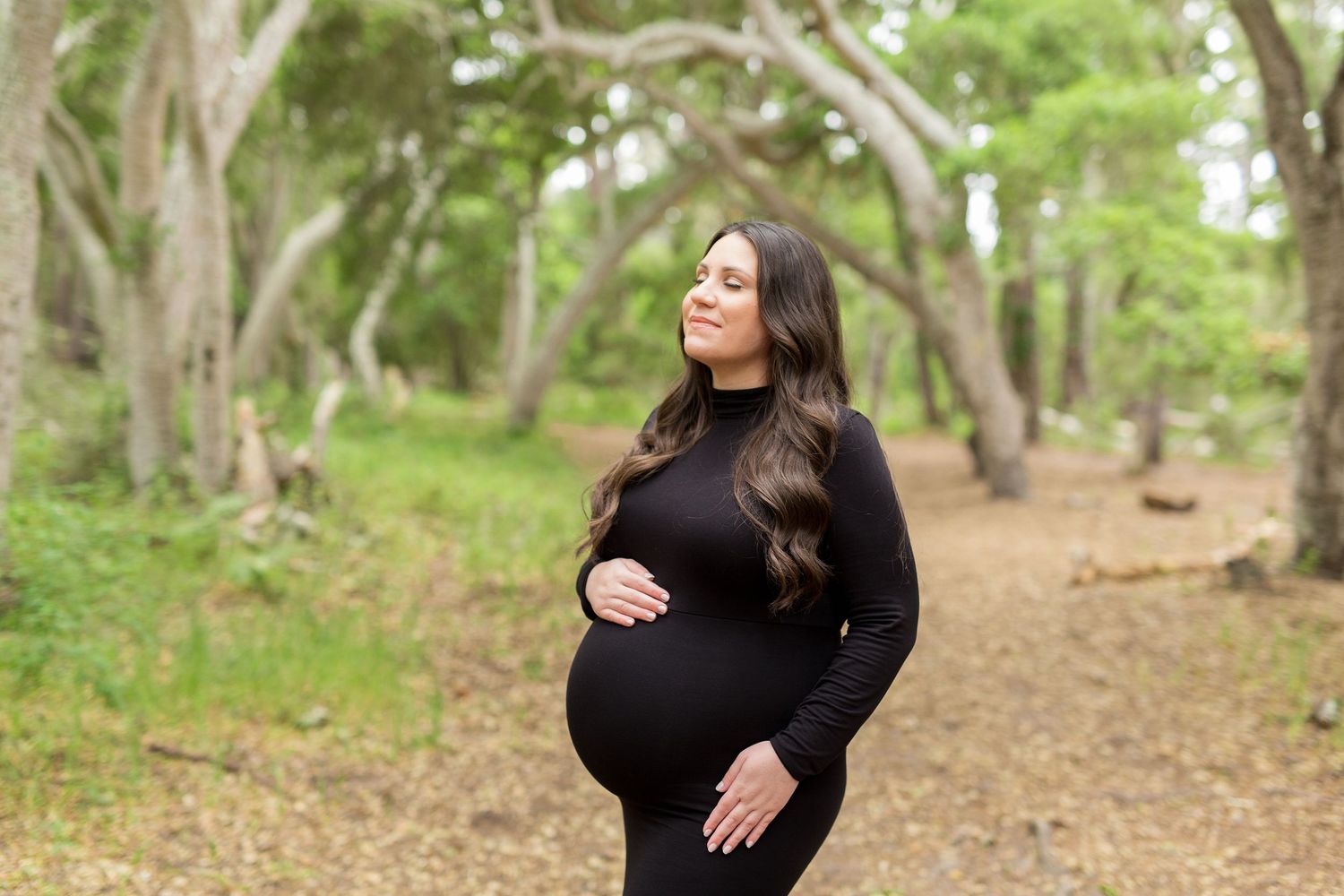 Woman in black dress standing in a wooded area during a maternity photoshoot.