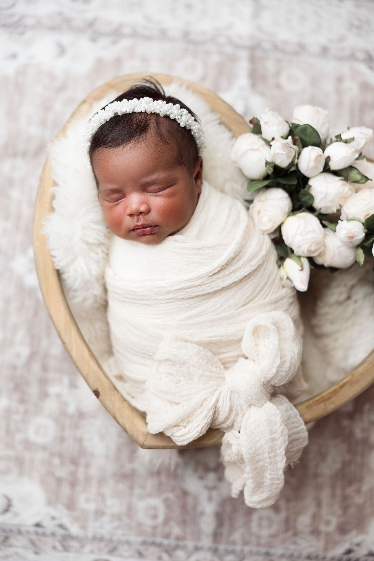 Studio newborn photo of baby girl swaddled in white wrap with floral headband, sleeping in a wooden heart bowl with white roses