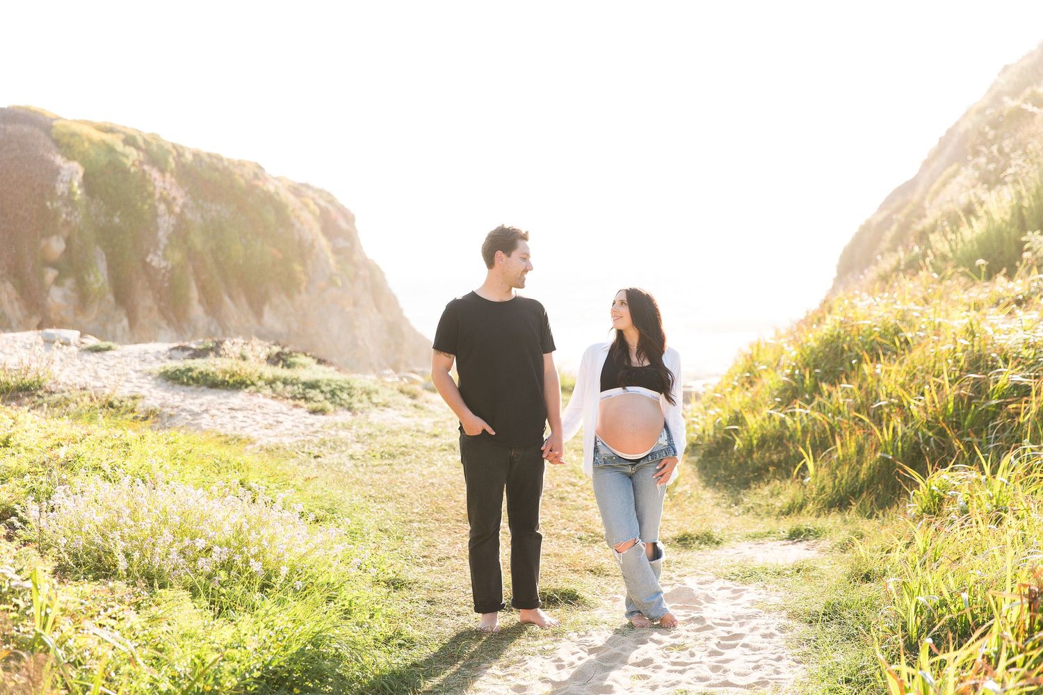 Couple walks along scenic coastal trail during golden hour maternity photography session.