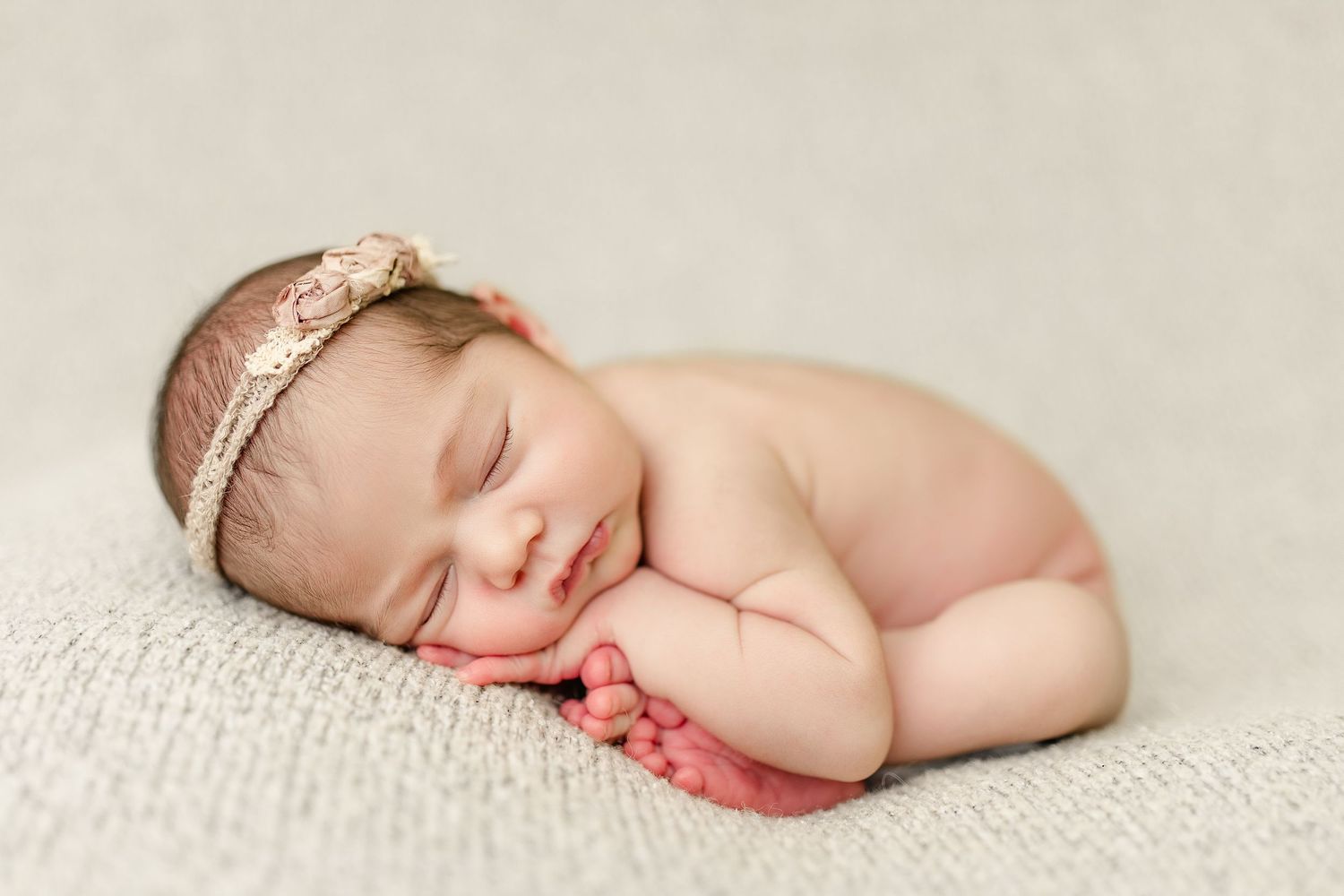 Peaceful sleeping newborn with delicate lace headband lying on textured cream blanket.