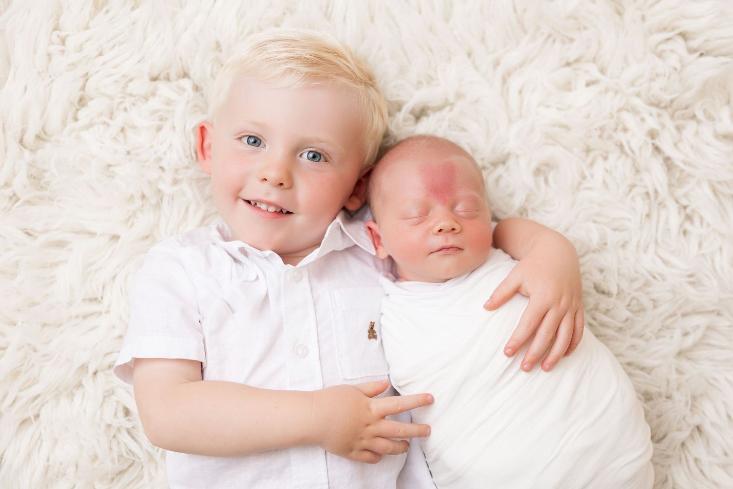Two siblings on white textured rug with newborn.