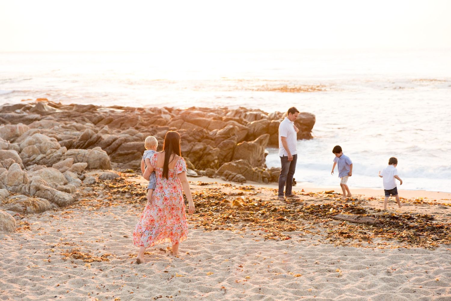 People walk along a sandy beach with rocky outcrops during sunset creating long shadows.