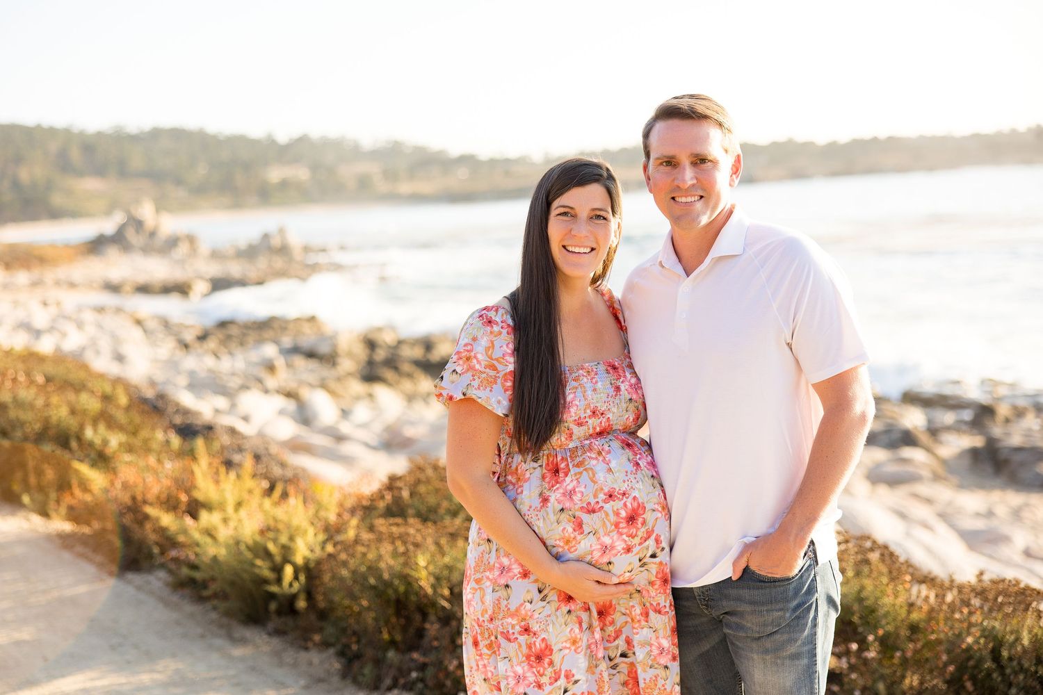 Expecting couple poses together on rocky coastal cliff during golden hour sunset.