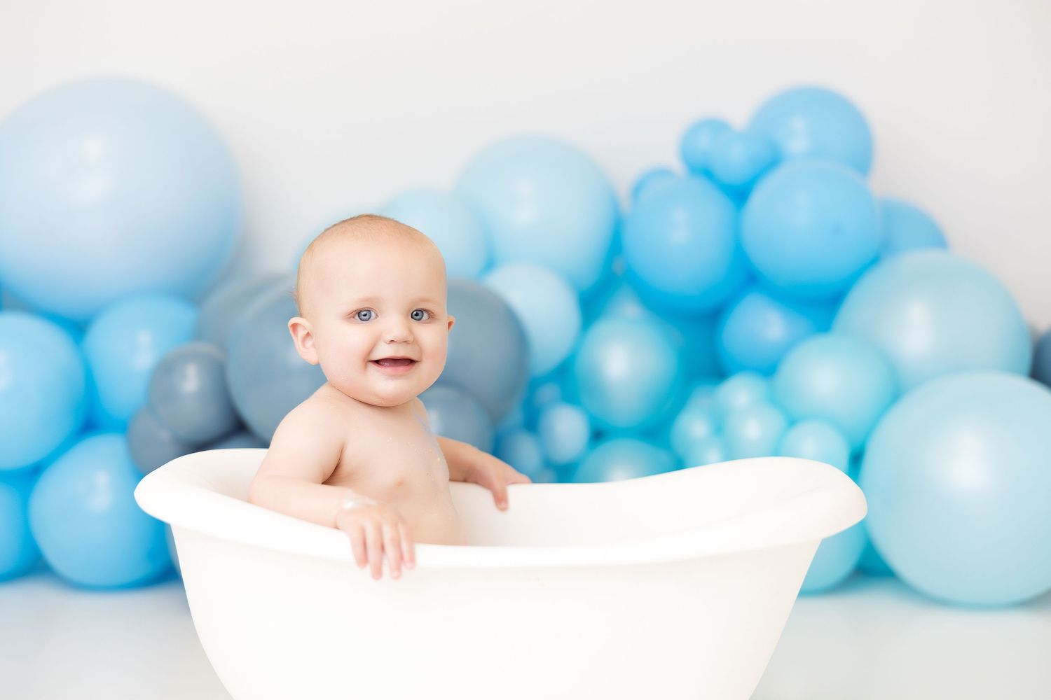 Smiling baby in white bathtub during cake smash cleanup with blue balloon decorations.