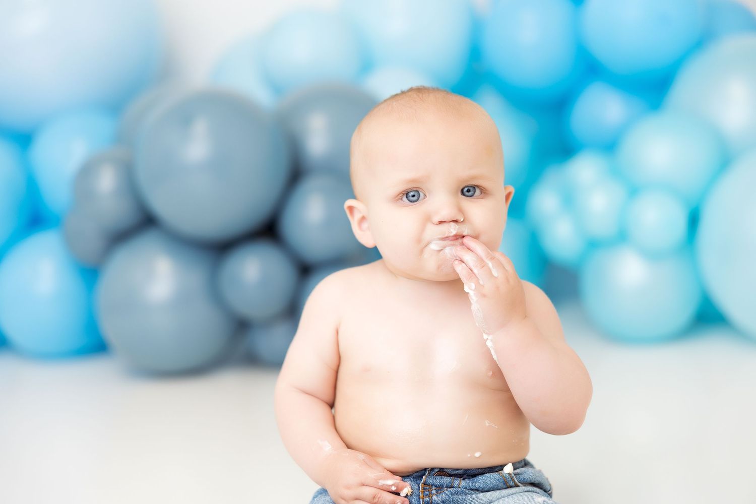 A baby wearing blue jeans sits and plays against a backdrop of blue and grey balloons.