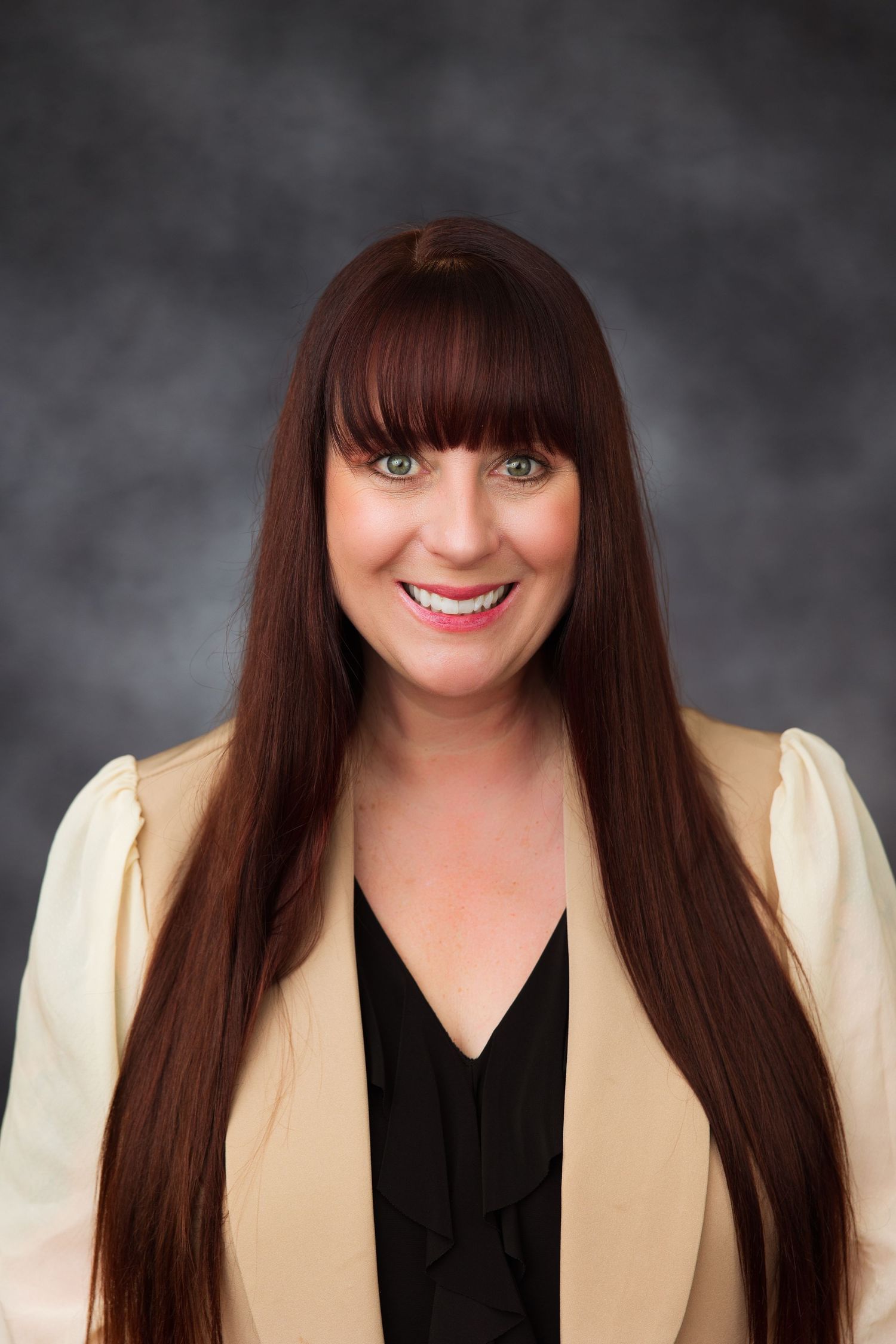 Professional headshot of a smiling woman with long auburn hair and bangs, wearing a cream blazer over a black top.