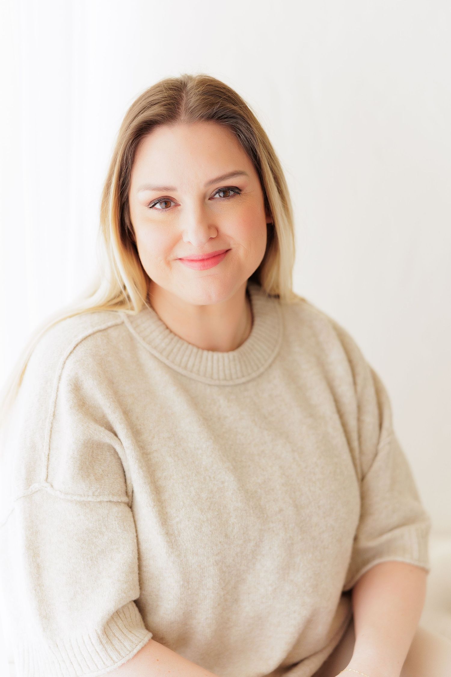 A smiling blonde woman wearing a beige knit sweater poses against a bright white background.