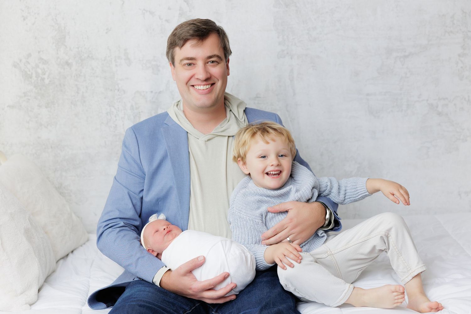 Father sitting with smiling toddler boy and newborn baby wrapped in white, all wearing light blue outfits.
