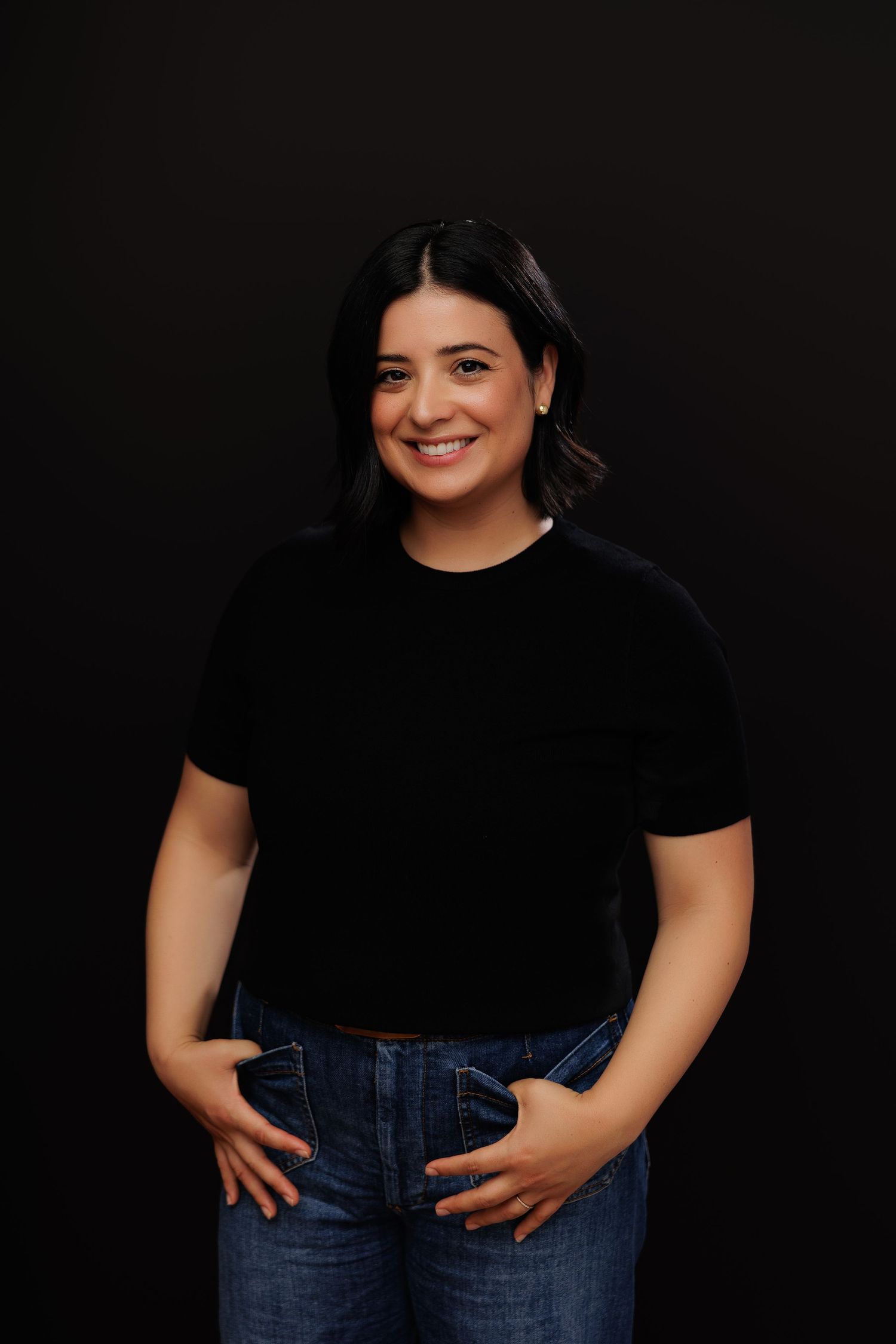 Smiling woman in black t-shirt and jeans posing confidently against a dark background.