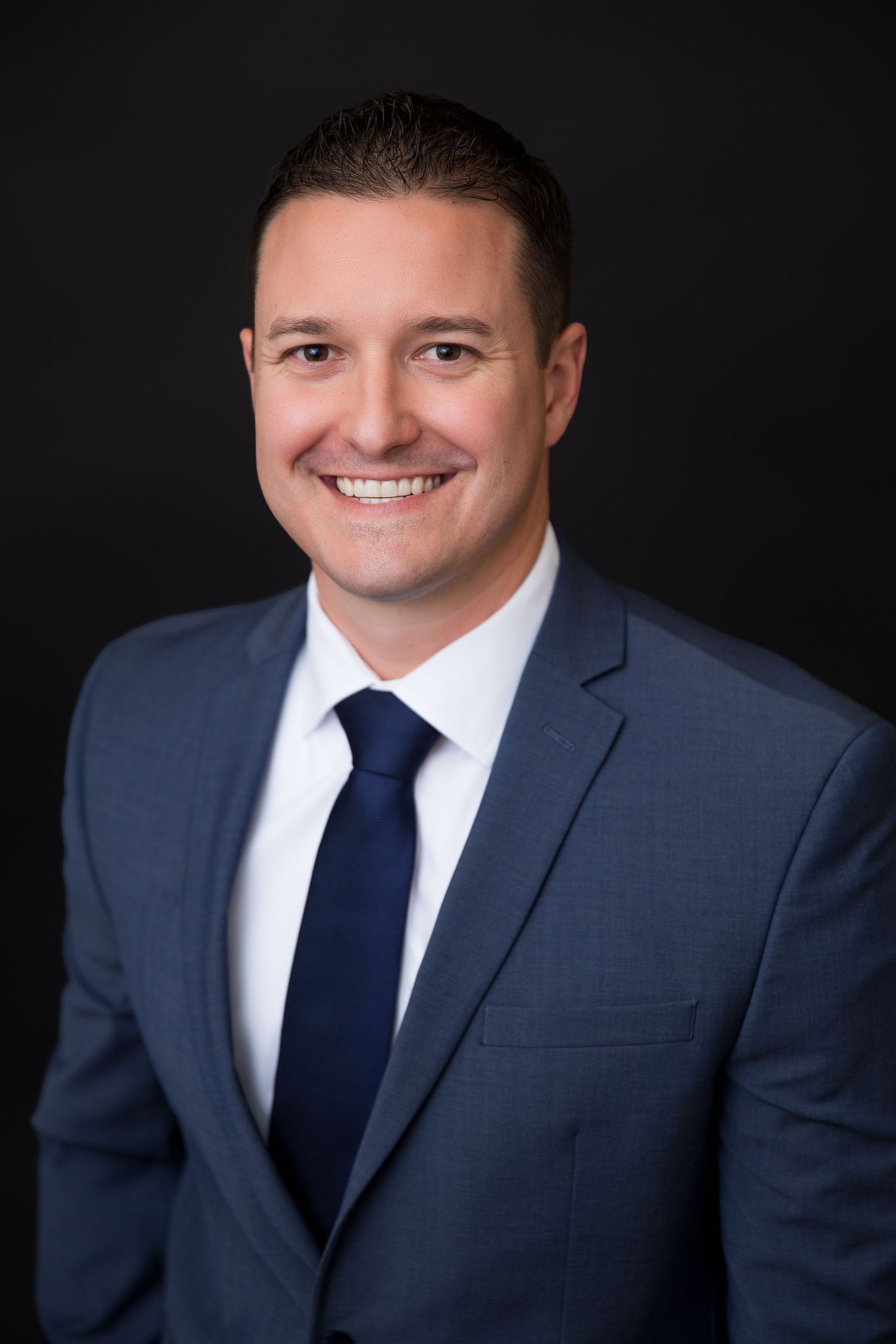 Professional headshot of a smiling man in a blue suit and navy tie against a dark background.