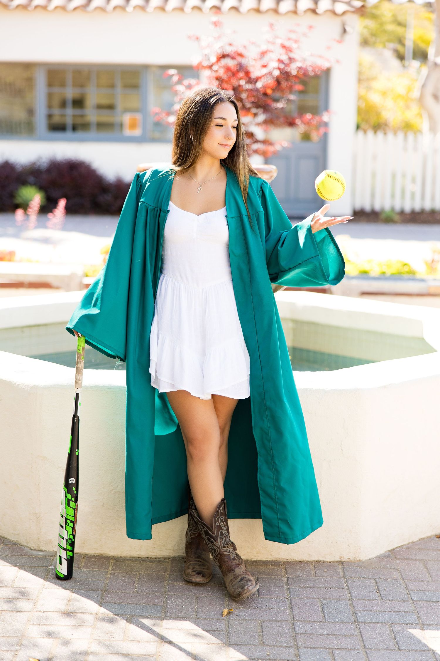 Young woman in green graduation gown, white dress, and cowboy boots holds tennis ball near fountain outdoors.