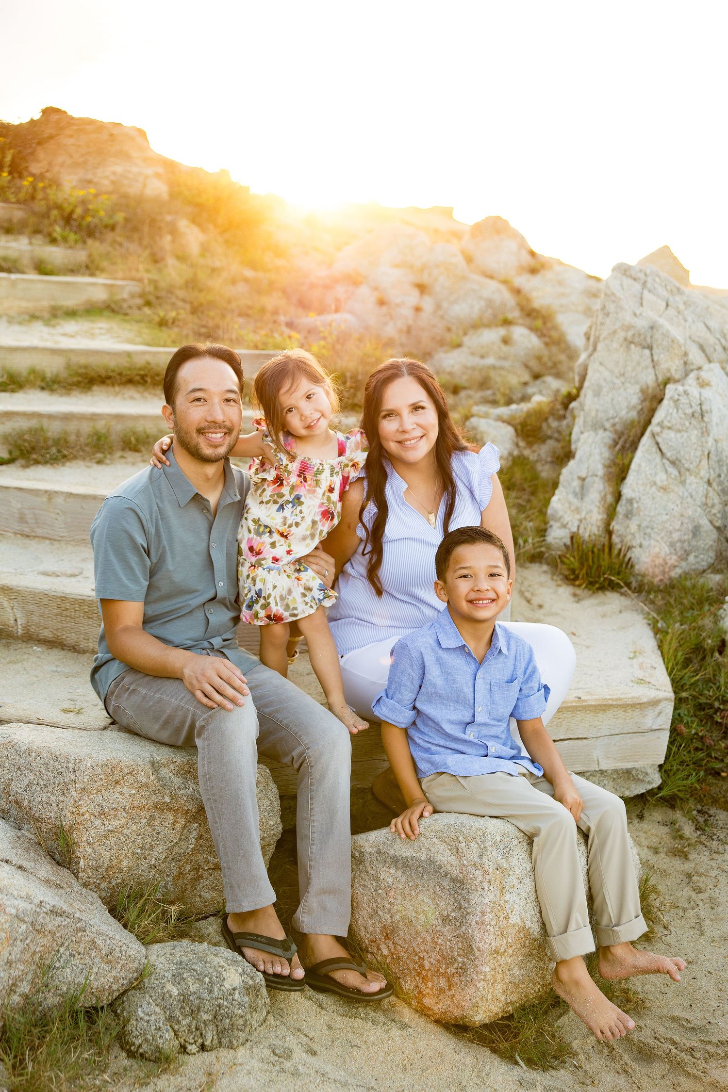 Family of four sitting on rocky steps at sunset, smiling warmly in casual blue and floral outfits at Asilomar Beach.