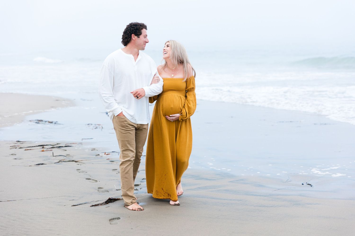 Couple strolling on beach during maternity session with woman in flowing yellow dress.