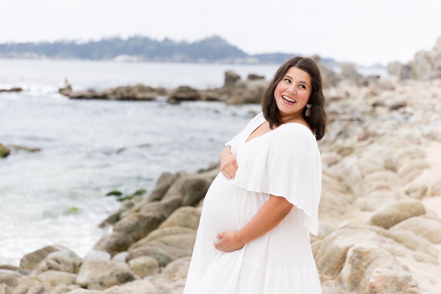 Expectant mother in white dress poses on rocky beach coastline during maternity photoshoot.