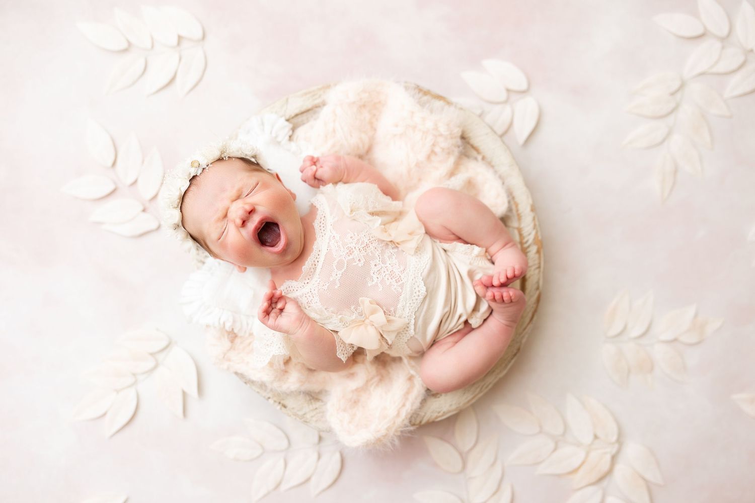 Yawning newborn in cream outfit lies on soft textured background with white paper flowers.