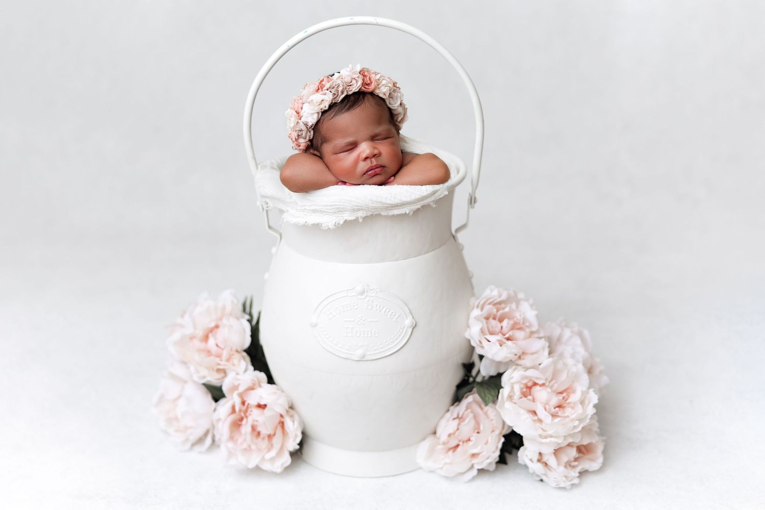 Newborn sleeping in white ceramic pot surrounded by pink roses against white backdrop.