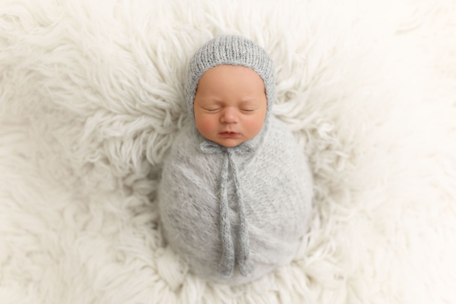Newborn wrapped in gray knit swaddle sleeping on white fur backdrop.