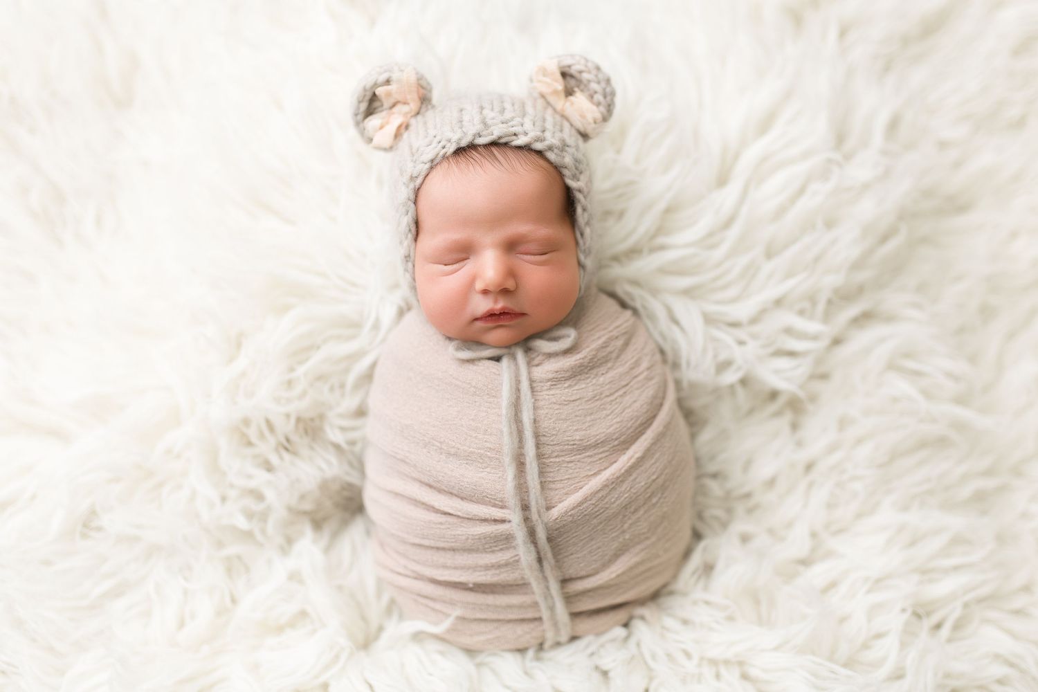 Sleeping baby wrapped in beige swaddle wearing cute bear ear hat on furry background.