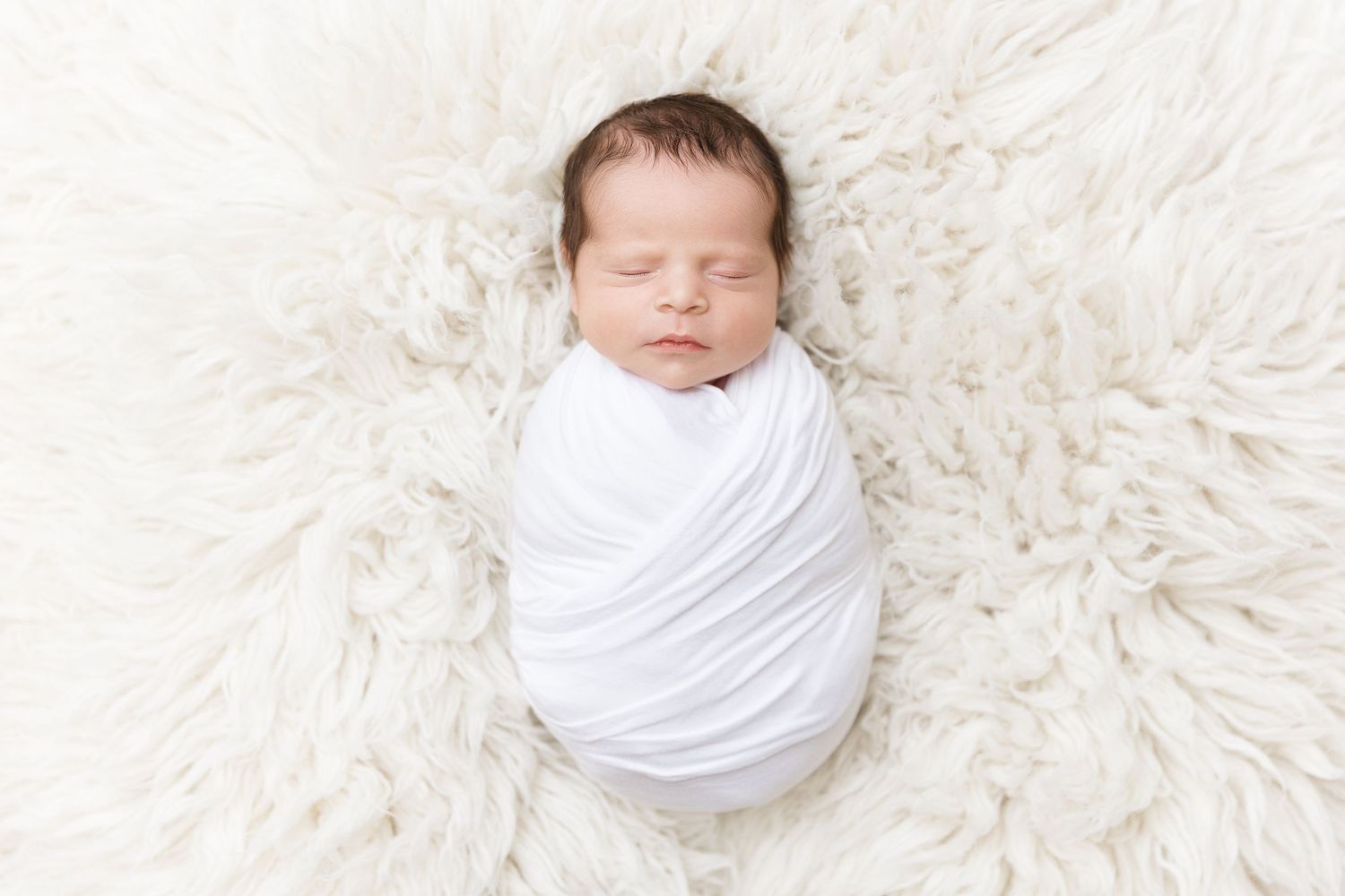 Newborn baby wrapped in white fabric sleeps on plush white fur rug.