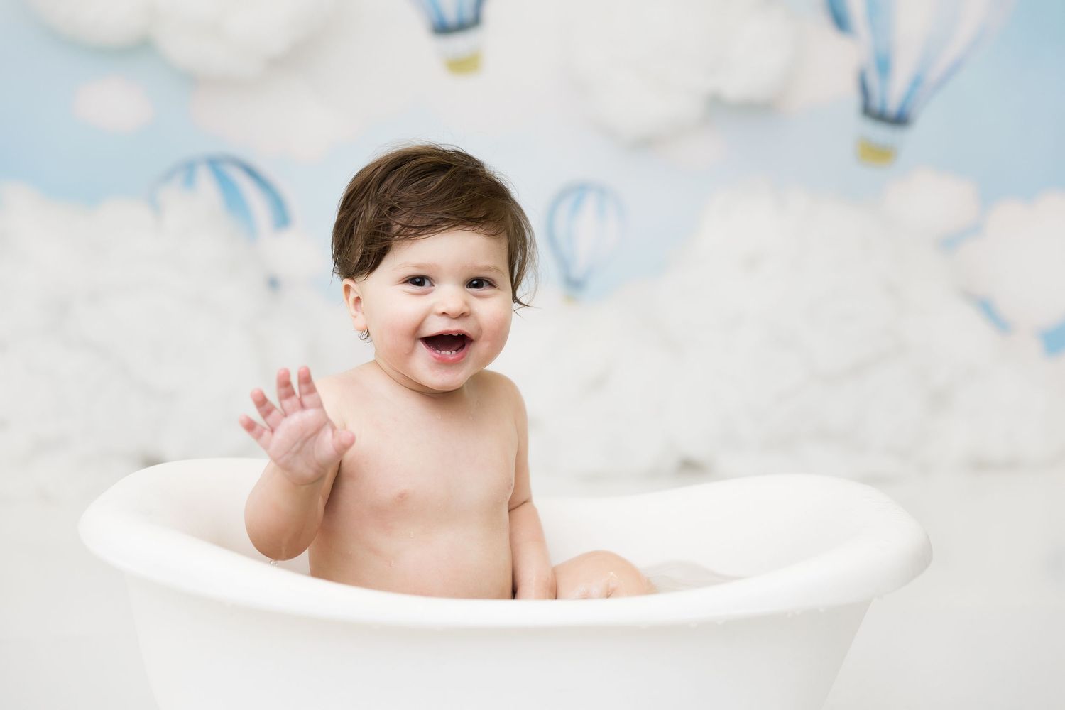 Joyful baby in white bathtub prop against cloud and hot air balloon themed background.