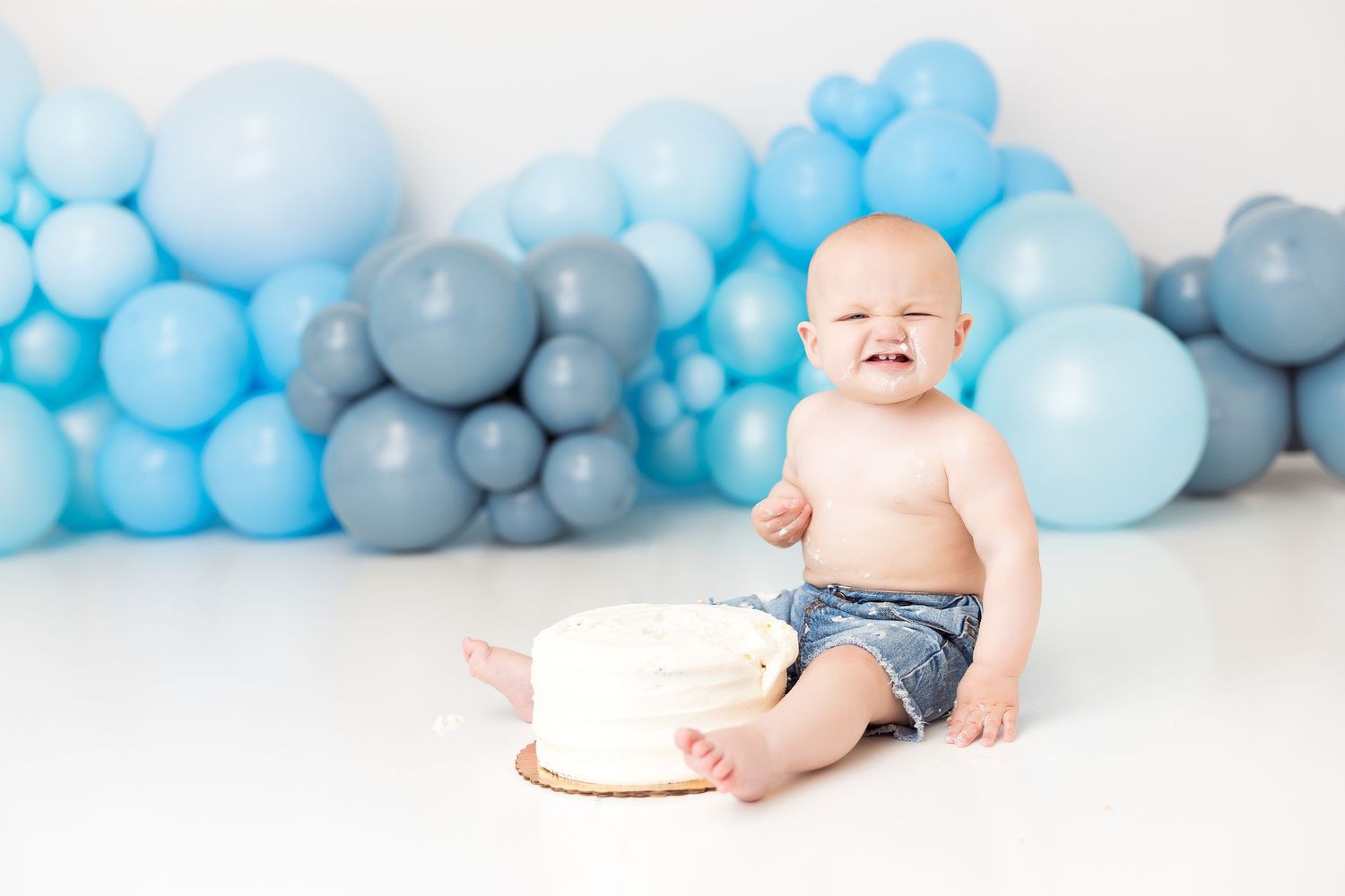 Baby in denim shorts crawls towards smash cake with blue balloon garland backdrop for first birthday celebration.