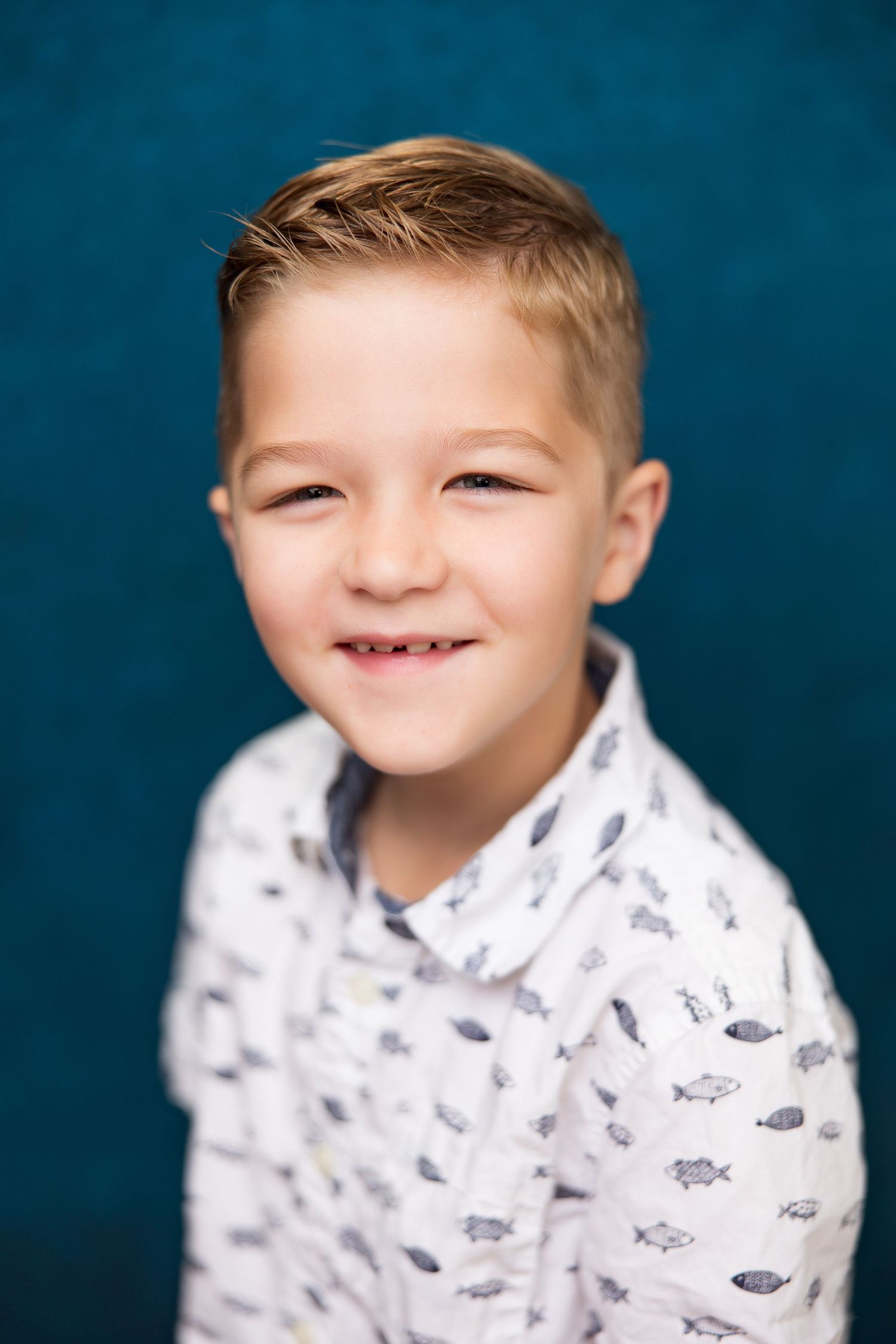 Smiling young boy with blonde hair wearing a white fish-print shirt, posed against a teal blue background.