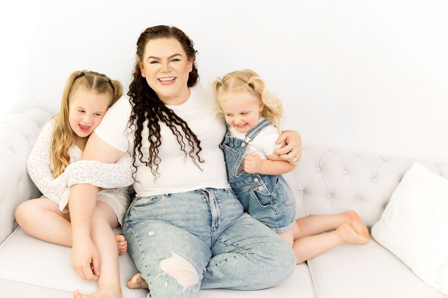 A woman with curly hair sits on a white couch smiling with two young blonde girls in a bright studio photo.