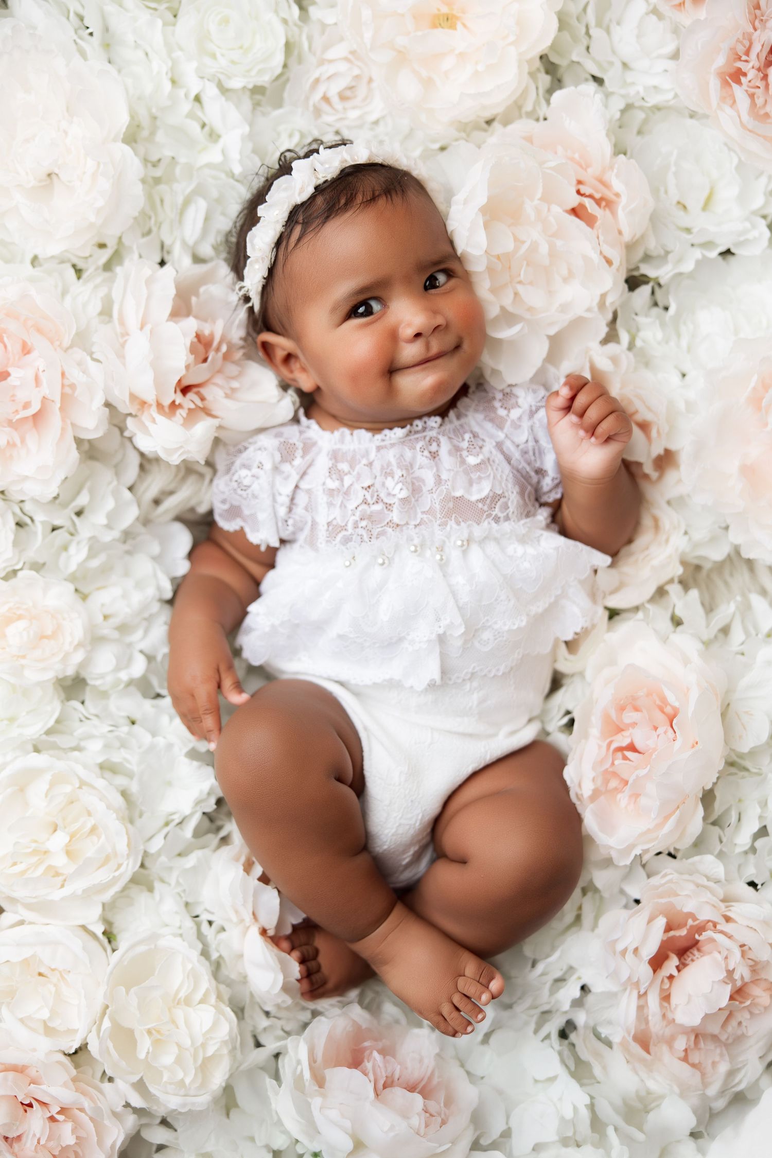Baby in white lace outfit lying on bed of soft pink and white roses.