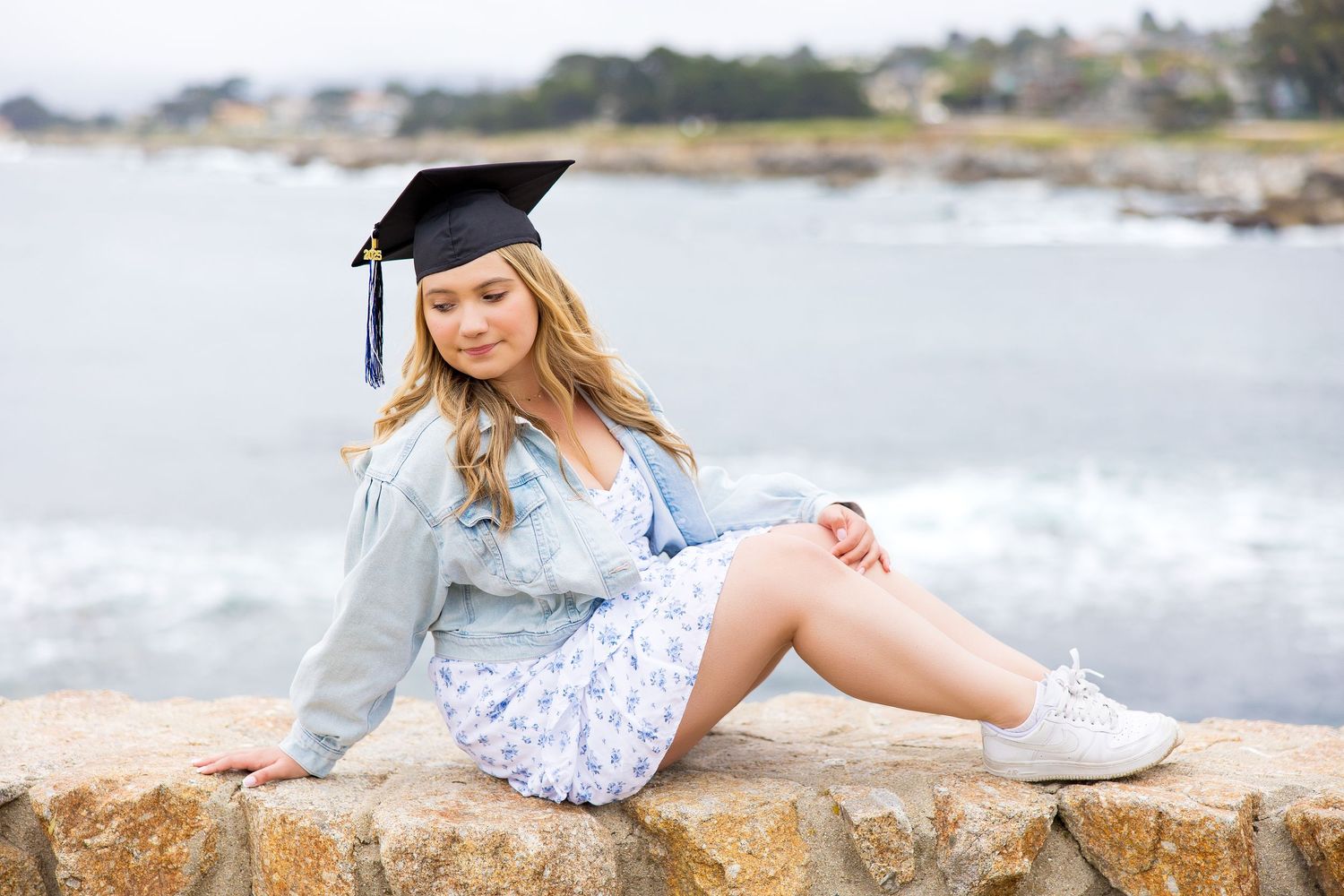 Young woman in graduation cap sitting on rocks by the ocean in a floral dress and denim jacket.