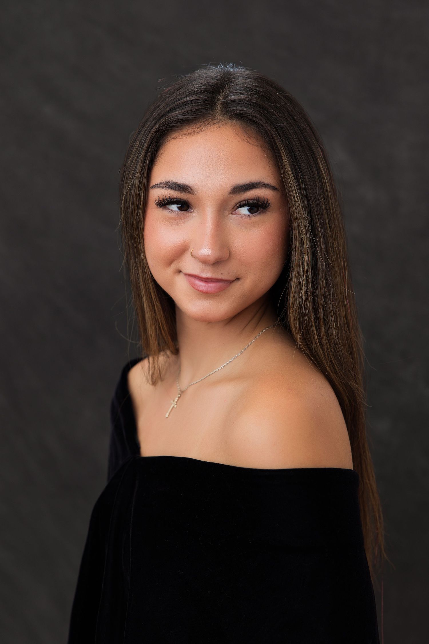 Young woman with long brown hair wearing a black off-shoulder top and delicate cross necklace poses for a portrait.