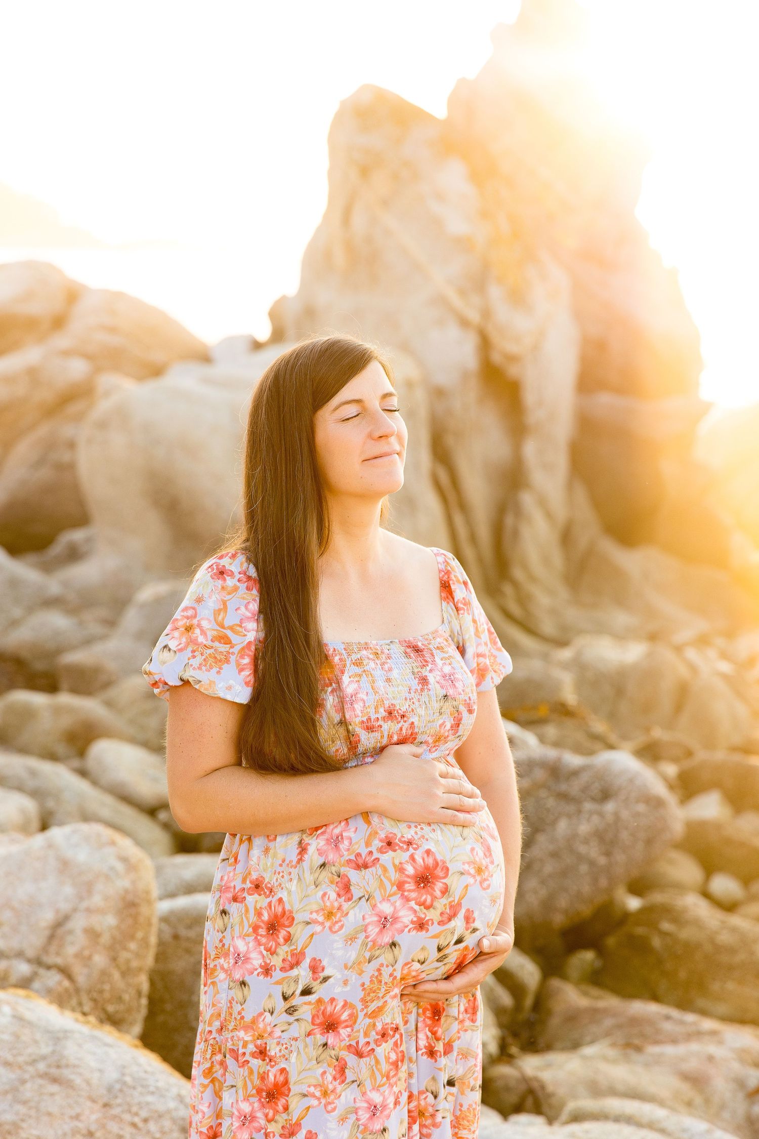 Woman in floral dress poses against rocky beach backdrop during golden hour maternity session.
