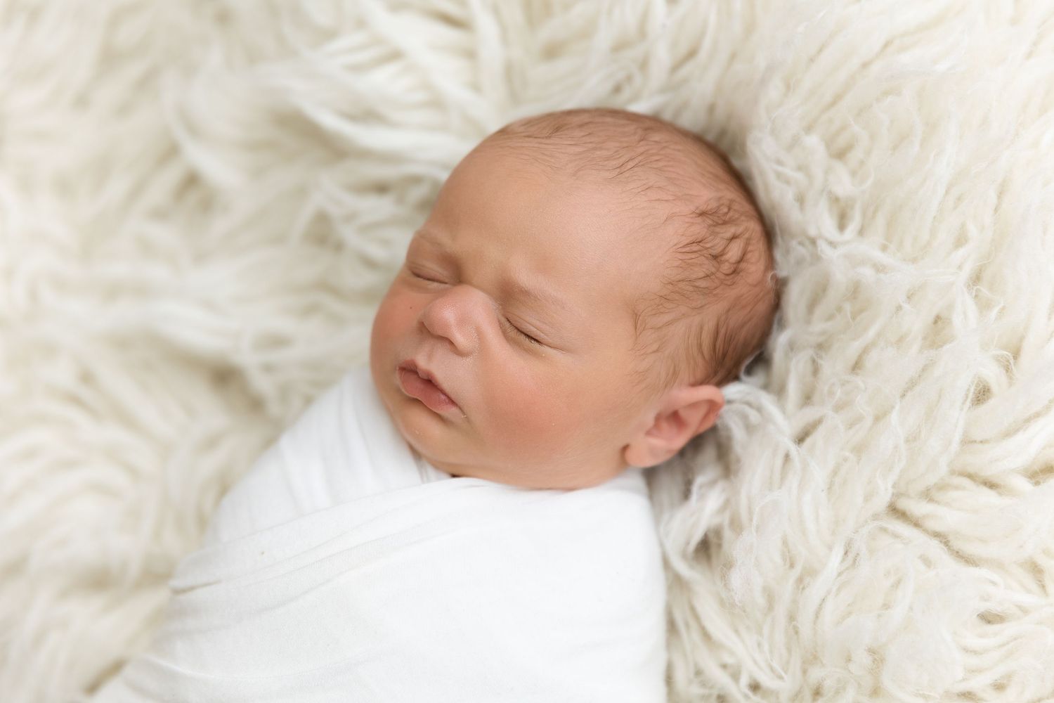Sleeping newborn baby wrapped in white blanket on fluffy white fur backdrop.