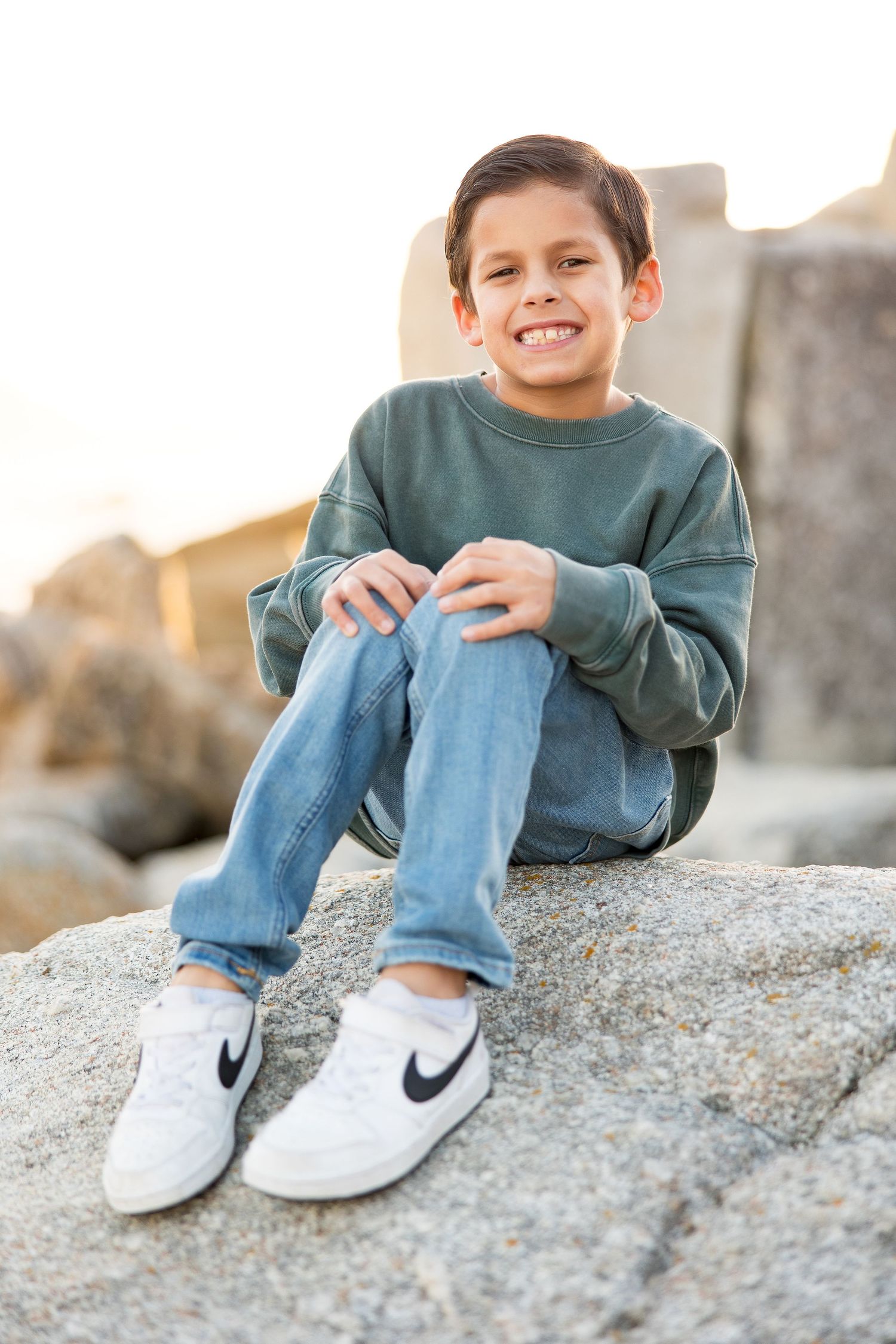 Child in green sweatshirt and jeans sitting on rocky terrain wearing Nike sneakers.