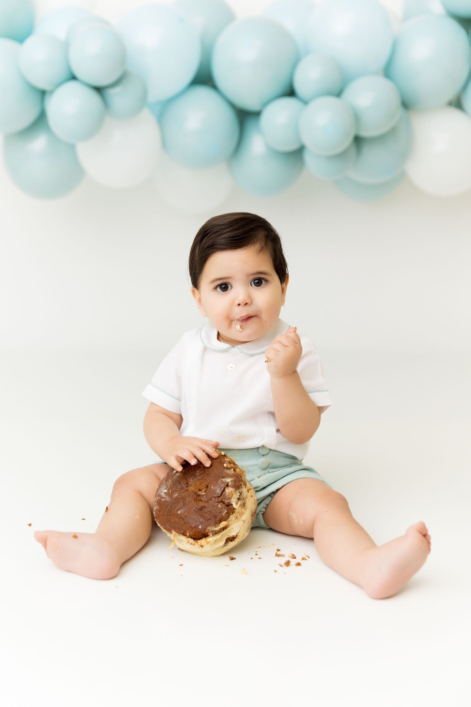 Baby in white shirt and mint shorts enjoys a chocolate dessert against blue balloon backdrop.