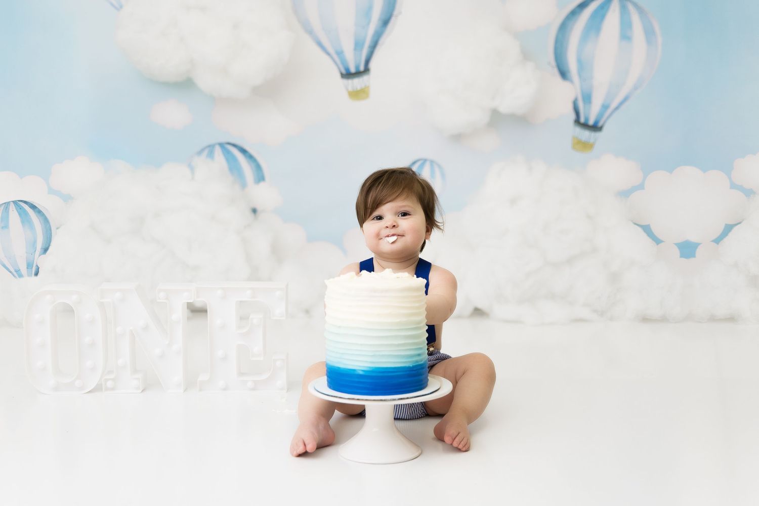 Baby with blue and white cake in hot air balloon themed photo setup.