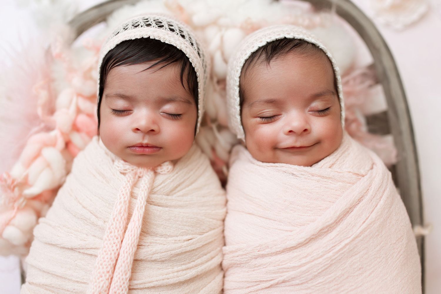 Twin babies wearing lace headbands sleep peacefully while wrapped in light pink swaddles.