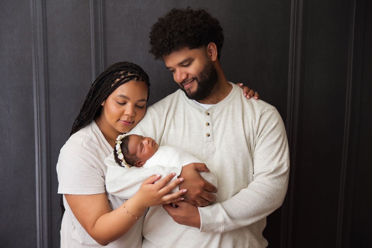 Parents holding newborn baby wrapped in white against dark background.