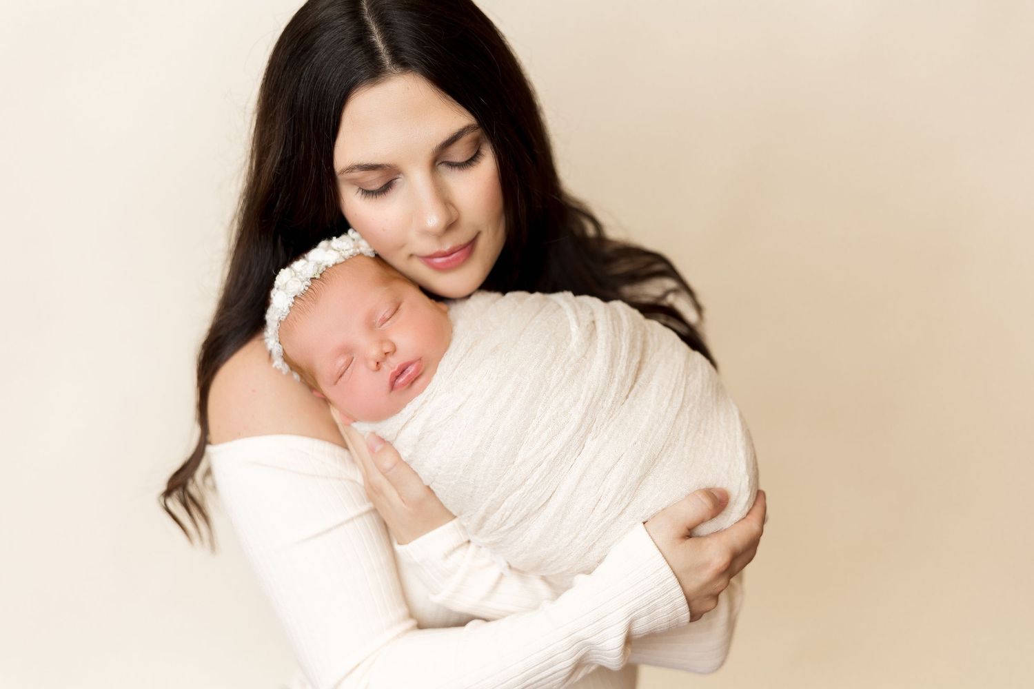 Parent wearing white cradles sleeping newborn swaddled in soft white fabric.