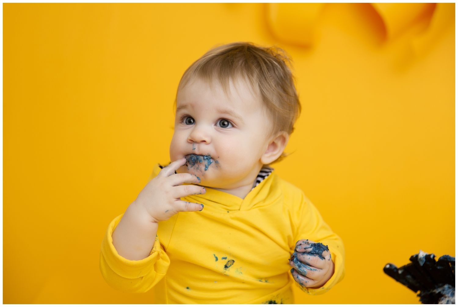 A small child in a yellow shirt sits against a yellow background while eating a snack.
