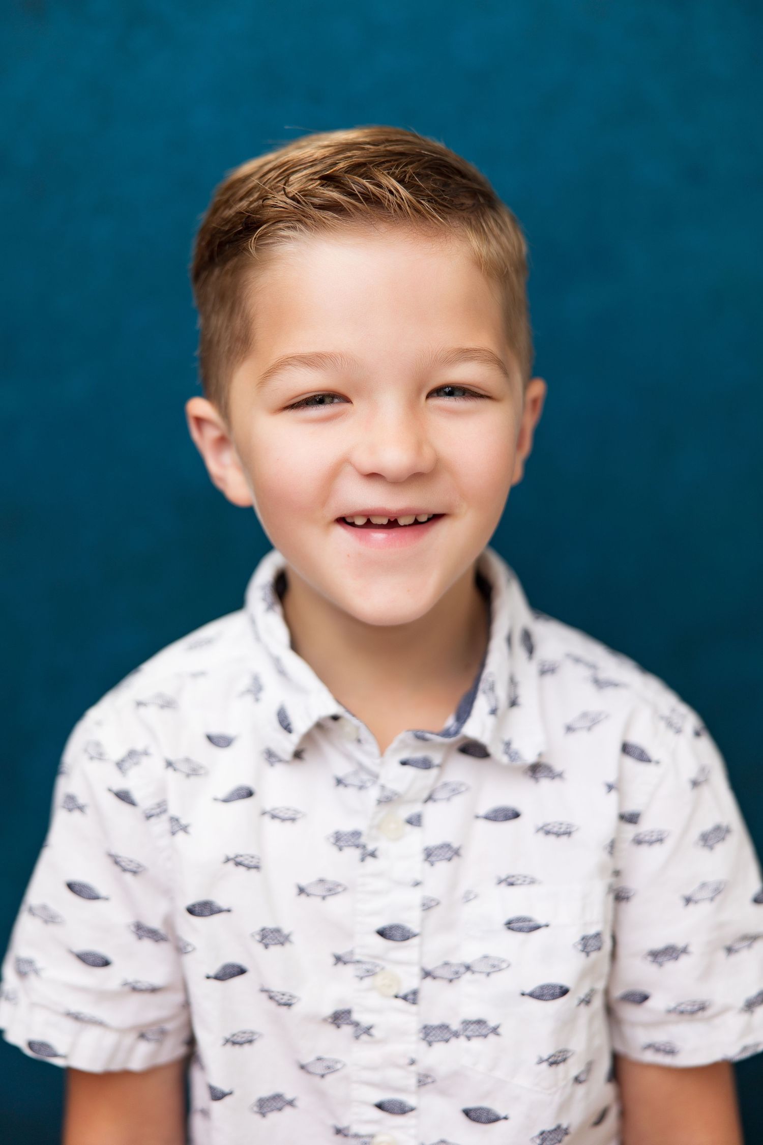 Smiling young boy with a missing front tooth wearing a white fish-print shirt against a teal background.