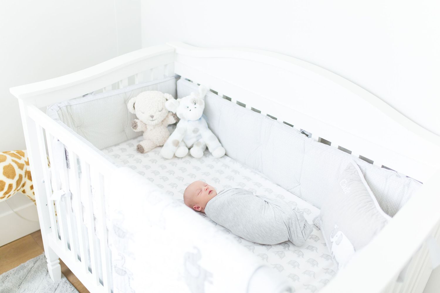 Baby in white sleepwear resting peacefully in a white crib with plush toys, lifestyle newborn session